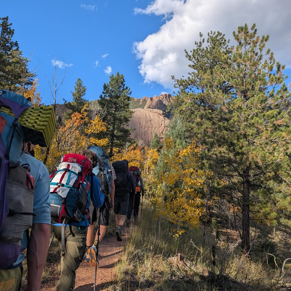 Students hiking a backcountry trail with full backpacking packs near a rock formation