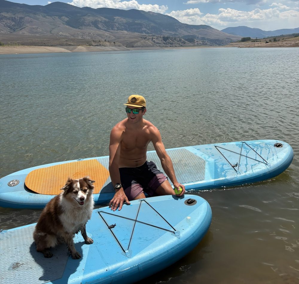 A student paddleboarding on a mountain lake with a dog on the board