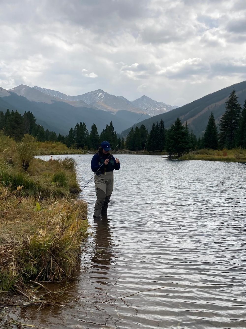 A student fly fishing in a mountain stream with peaks in the background